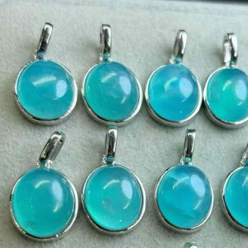 May include: A selection of oval pendants featuring a vibrant, translucent teal gemstone set in silver-coloured metal. Each pendant has a small loop at the top for a chain. The pendants are closely arranged, indicating a jewellery collection.