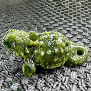 Op de afbeelding: Een gedetailleerde, groene jade-snijwerk van een hagedis. De sculptuur heeft een textuur met een hobbelig, schubachtig patroon. De hagedis is geposeerd met zijn poten ingetrokken en staart opgerold.
