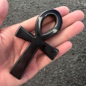 May include: A polished, smooth, black ankh symbol carved from stone. The object features the classic Egyptian cross shape with a looped top and three extending arms, displaying a reflective, glossy surface texture throughout the entire piece.