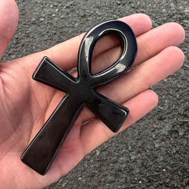 May include: A polished, smooth, black ankh symbol carved from stone. The object features the classic Egyptian cross shape with a looped top and three extending arms, displaying a reflective, glossy surface texture throughout the entire piece.