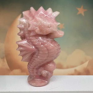 May include: A pink seahorse figurine carved from a polished stone. The seahorse has a spiky dorsal fin, detailed fins, and a curled tail. The figurine is a light pink color and has a glossy finish.