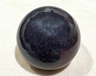 May include: A polished, spherical gemstone with a deep blue hue and sparkling inclusions. The sphere has a smooth, reflective surface, and the inclusions create a starry effect. The gemstone is set against a neutral background.