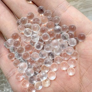 May include: A handful of clear, spherical glass beads with a slightly pinkish hue. The beads are smooth and translucent, reflecting light. The image shows a close-up view of the beads, suggesting they are small and delicate. The beads are likely used for crafting or decorative purposes.