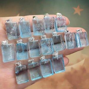 May include: A collection of rectangular, light blue gemstone pendants with silver-colored metal caps. The pendants are transparent, revealing internal patterns. The image shows multiple pendants, each with a loop for a chain.