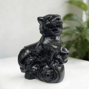 Könnte beinhalten: Eine massive schwarze Tigerfigur in sitzender Position mit geöffnetem Maul, das Zähne zeigt. Der Tiger sitzt auf einem runden Sockel, der mit komplizierten, geprägten kreisförmigen Mustern und dekorativen Schnörkeln am unteren Rand versehen ist.