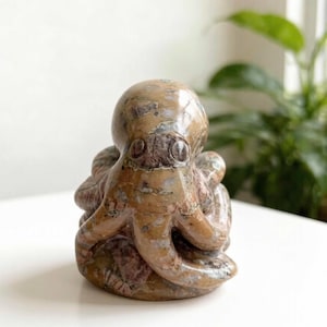 Peut inclure: Une sculpture en pierre polie représentant une pieuvre avec une finition lisse et brillante. L'objet présente un motif tacheté dans des tons de beige, de brun et de gris sur toute sa tête arrondie et ses tentacules enroulés.