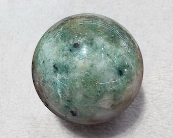 May include: A spherical decorative object with a mottled appearance. The sphere features a mix of green, white, and brown hues, with darker spots scattered across its surface. The overall effect is a natural, earthy aesthetic.