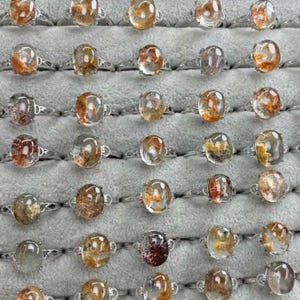 May include: A display of silver rings, each featuring an oval-cut, clear gemstone with internal patterns in shades of orange, brown, and yellow. The rings are neatly arranged on a grey surface, highlighting the different designs.
