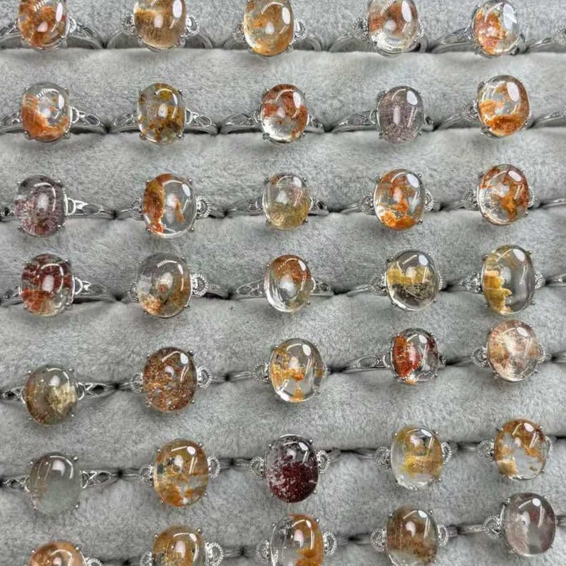 May include: A display of silver rings, each featuring an oval-cut, clear gemstone with internal patterns in shades of orange, brown, and yellow. The rings are neatly arranged on a grey surface, highlighting the different designs.