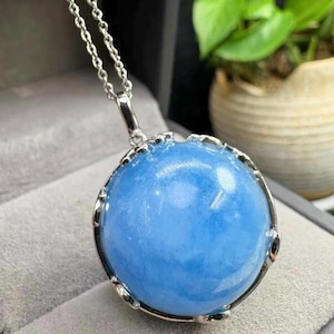 May include: A silver necklace featuring a large, round pendant with a light blue, translucent stone. The stone is encased in a decorative silver setting and hangs from a delicate silver chain. The pendant has a smooth, polished surface.