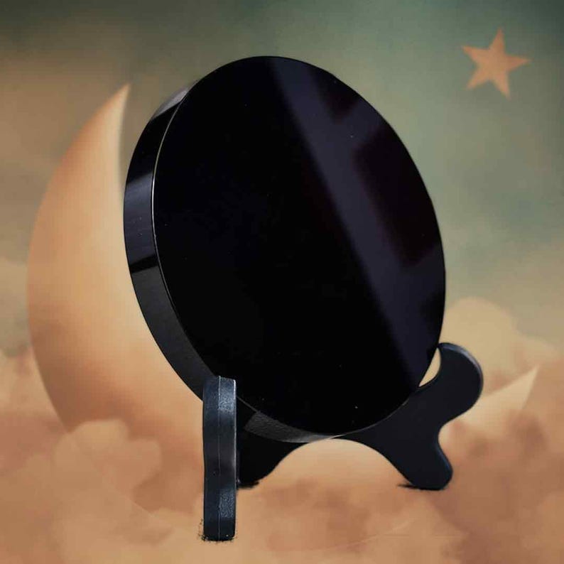 May include: A circular, black, polished obsidian scrying mirror on a black stand. The mirror is set against a backdrop of a crescent moon, clouds, and a star. The stand has a decorative, curved design.