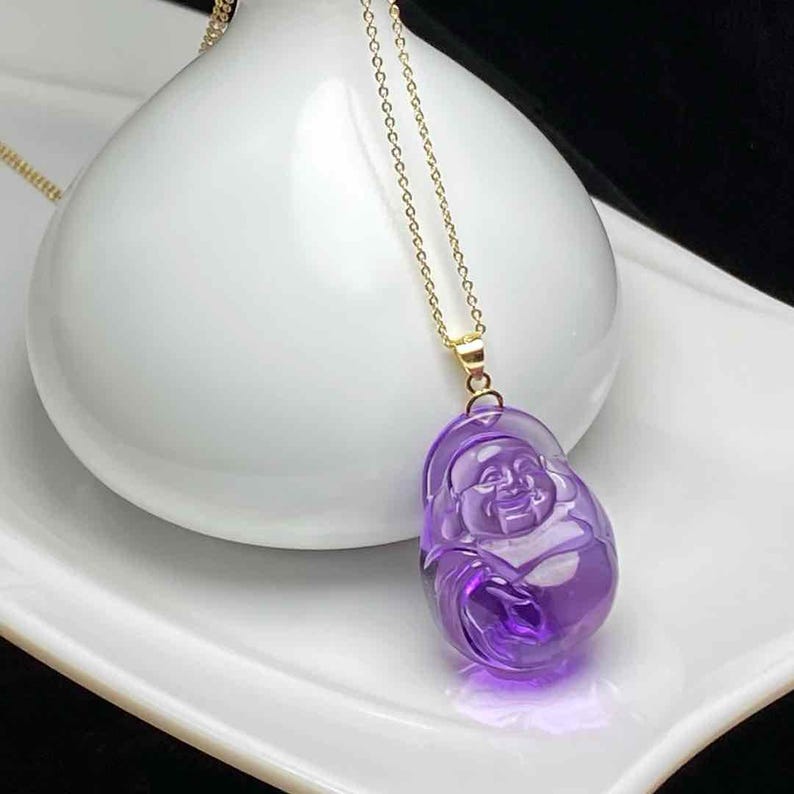 May include: A purple amethyst pendant necklace featuring a carved laughing Buddha figure. The pendant hangs from a delicate gold chain. The Buddha is a vibrant shade of purple, with intricate details.