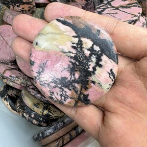May include: A polished, oval-shaped gemstone with a pink base color, featuring black and white veining patterns. The stone is held in a hand, with other similar stones visible in the background. The overall aesthetic is natural and earthy.