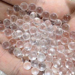 May include: A collection of clear, spherical glass beads. The beads are transparent with a slight pinkish hue, and are held in a hand. The beads are smooth and reflect light, creating a sparkling effect. The overall theme is of decorative elements or craft supplies.