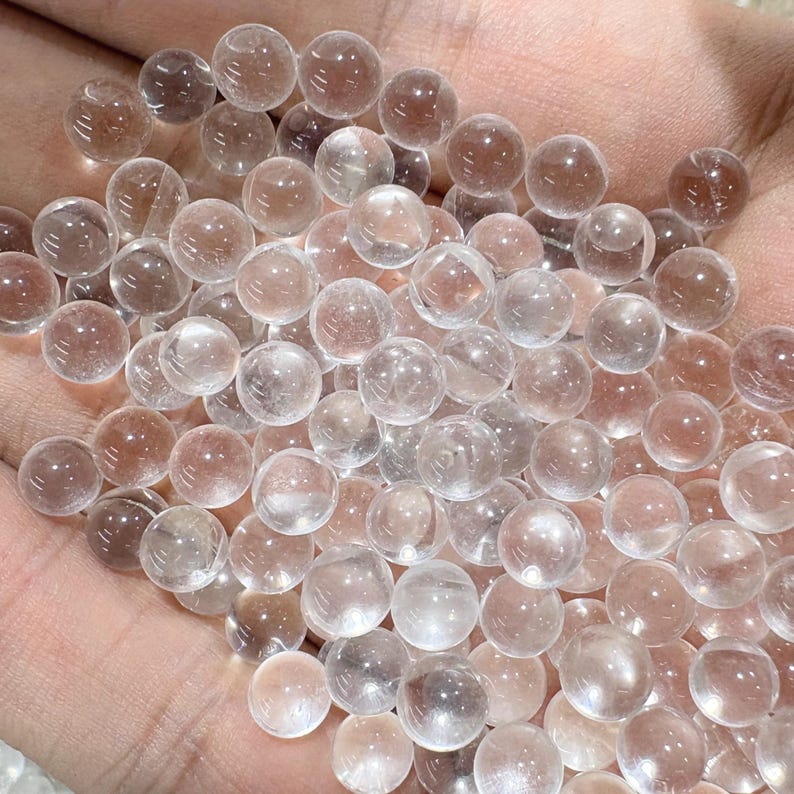 May include: A collection of clear, spherical glass beads. The beads are transparent with a slight pinkish hue, and are held in a hand. The beads are smooth and reflect light, creating a sparkling effect. The overall theme is of decorative elements or craft supplies.
