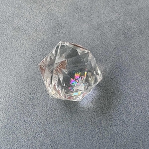 Puede incluir: Una piedra de cristal transparente y facetada con forma geométrica. La superficie translúcida presenta bordes angulares afilados y fracturas internas que crean una vibrante refracción de luz de colores arcoíris en el centro.
