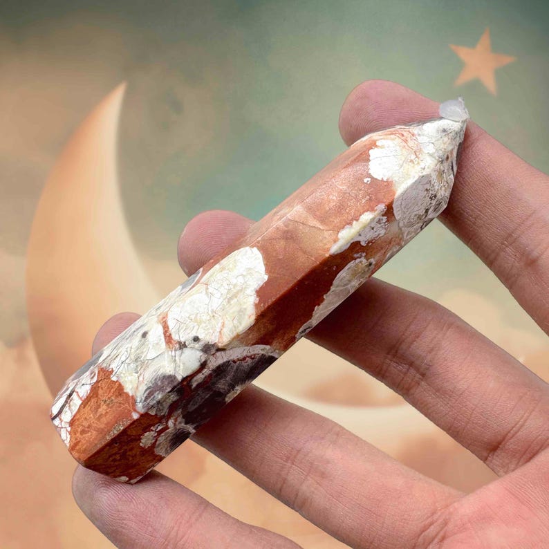 May include: A reddish-brown and white crystal point held in a hand. The crystal has a hexagonal shape and a polished surface. The background features a crescent moon and a star.