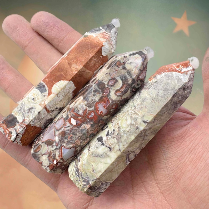 May include: Three polished gemstone obelisks in shades of brown, red, and cream are held in a hand. Each obelisk has a faceted, pointed top and a unique pattern of natural markings. The gemstones are approximately 5 inches long.