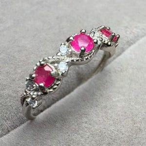 May include: A silver ring featuring three oval-cut, vibrant pink gemstones and smaller clear stones. The ring has a delicate, ornate design with a polished finish. The gemstones are set in a row, creating a continuous pattern.
