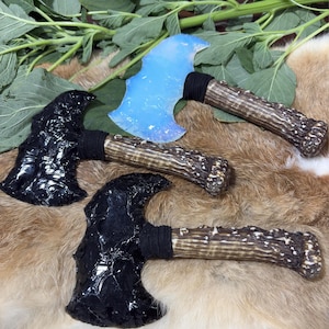 May include: Three decorative axes with obsidian and opalescent blades. The handles are made of antler and wrapped with black fabric. The axes are displayed on a fur-like surface with green foliage.
