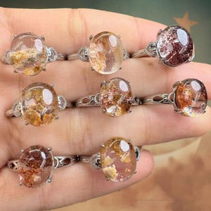 May include: A collection of gemstone rings featuring oval-cut stones in diverse colours, such as clear, brown, and reddish tones. The rings have silver-coloured bands with decorative detailing around the stone settings. The gemstones have inclusions.