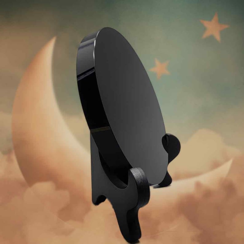 May include: A black, circular decorative stand with a glossy finish. The stand has a curved base and a round top, designed to hold a plate or other display item. The background features a crescent moon and stars.