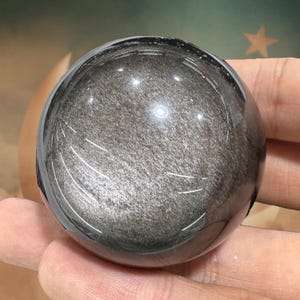 May include: A polished, spherical obsidian gemstone. The sphere is a dark, reflective gray with a shimmering, metallic appearance. The surface has a smooth, glossy finish, and the overall shape is perfectly round.