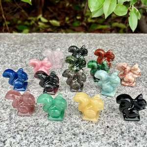 Puede incluir: Una colección de pequeñas figuras de ardillas talladas en varios colores, incluyendo azul, rosa, verde y amarillo. Las figuras están hechas de piedra pulida y se exhiben sobre una superficie gris.