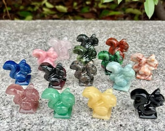 May include: A collection of small, carved squirrel figurines in various colors, including blue, pink, green, and yellow. The figurines are made of polished stone and are displayed on a gray surface.