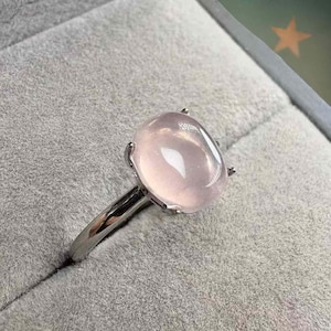 May include: A silver ring featuring a large, oval-shaped, pale pink gemstone. The gemstone is set with four prongs, and the ring band is smooth and polished. The ring is displayed on a gray surface.