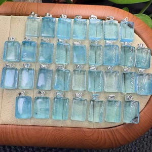 May include: A collection of rectangular, light blue pendants with silver-colored bails. The pendants are arranged on a tan surface, showcasing their translucent quality and variations in color. The pendants are likely for necklaces or other jewelry.