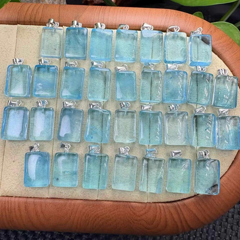 May include: A collection of rectangular, light blue pendants with silver-colored bails. The pendants are arranged on a tan surface, showcasing their translucent quality and variations in color. The pendants are likely for necklaces or other jewelry.