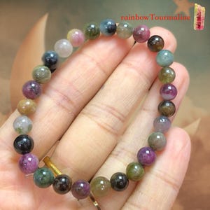 May include: A multi-colored tourmaline bead bracelet. The bracelet features a variety of colors, including pink, green, and black. The beads are round and strung together. The text "rainbowTourmaline" is visible in the image.