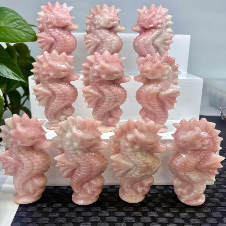 May include: A collection of pink seahorse figurines. Each figurine is carved from a light pink stone, with detailed fins, tails, and spiky crests. The seahorses are arranged on a white and black surface, showcasing their intricate design and color variations.