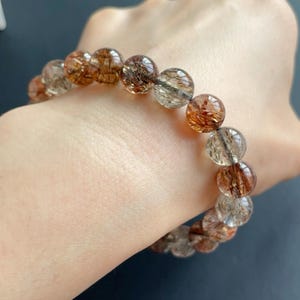 May include: A beaded bracelet featuring translucent, spherical beads with internal reddish-brown inclusions. The beads are strung together, creating a bracelet. The bracelet is held against a neutral background.