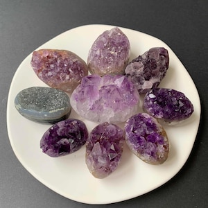 May include: A collection of eight small amethyst crystal clusters and one smooth grey agate stone arranged in a circular pattern. The amethyst pieces feature varying shades of purple, ranging from light lavender to deep violet, with jagged, crystalline surfaces.