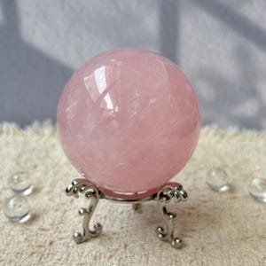 Pode incluir: Uma esfera de quartzo rosa polido, repousando sobre um suporte de metal prateado ornamentado. A esfera é de cor rosa suave e reflete a luz. O suporte tem um design decorativo enrolado. Esferas de vidro transparente espalhadas ao redor da base.