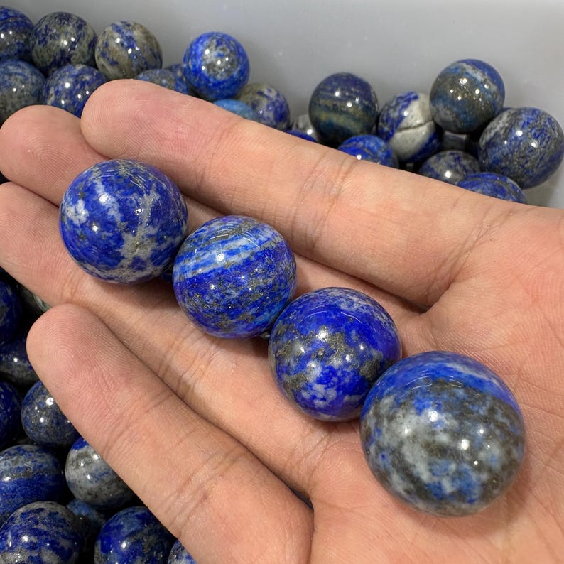 May include: Several polished lapis lazuli spheres, each a deep blue with grey and white streaks. The spheres, of varying sizes, are held in a hand, highlighting their smooth, reflective surfaces. These decorative spheres are ideal for home decor.