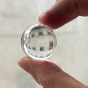 Op de afbeelding: Een heldere, bolvormige glazen lens die tussen twee vingers wordt gehouden. De lens reflecteert een vervormd beeld van een gebouw met ramen. Het glas is glad en transparant, met een lichte kromming.