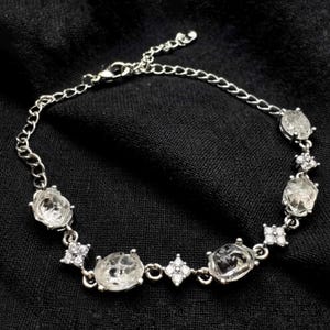 Op de afbeelding: Een zilverkleurige armband met heldere, ovaalvormige edelstenen en kleine, ronde accentstenen. De armband is geplaatst tegen een zwarte achtergrond, waardoor de sprankelende details worden benadrukt. De ketting is delicaat en verstelbaar.