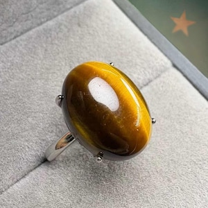 May include: A polished tiger's eye gemstone ring. The oval-shaped stone is a mix of brown and gold hues, set in a silver band. The ring is displayed on a gray surface.