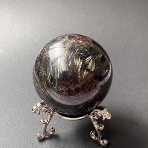 Puede incluir: Una piedra esférica pulida con un color base púrpura oscuro y negro, con prominentes inclusiones brillantes en forma de aguja de color marrón dorado. La esfera descansa sobre un soporte decorativo de trípode de metal plateado.