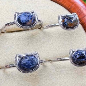 May include: Four silver rings with a cat-shaped setting. Each ring features a blue and brown gemstone in an oval shape. The cat-shaped setting has a beaded border. The rings are displayed on a light beige surface.