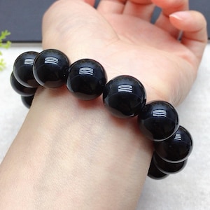 Puede incluir: Un brazalete de cuentas negras con cuentas grandes y redondas. Las cuentas tienen un acabado brillante y reflejan la luz. El brazalete se lleva en una muñeca, mostrando el tamaño y el diseño de la joya. El brazalete es de color oscuro y neutro.