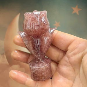 May include: A carved rose sculpture made of translucent, reddish-pink stone. The rose is in full bloom, with detailed petals and a stem. The sculpture is held in a hand, with a soft background of a moon and stars.