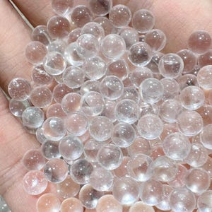 May include: A collection of clear, spherical crystal balls. The spheres vary slightly in color, with some appearing more transparent and others having a subtle pinkish hue. The balls are smooth and appear to be made of glass or crystal.