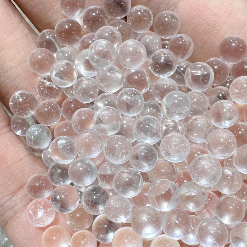 May include: A collection of clear, spherical crystal balls. The spheres vary slightly in color, with some appearing more transparent and others having a subtle pinkish hue. The balls are smooth and appear to be made of glass or crystal.