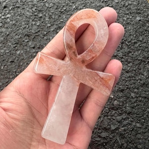 May include: An ankh symbol carved from translucent quartz with visible reddish-brown mineral inclusions throughout the stone. The object features a smooth, polished surface with a rounded top loop and a vertical base, showcasing natural crystalline patterns.
