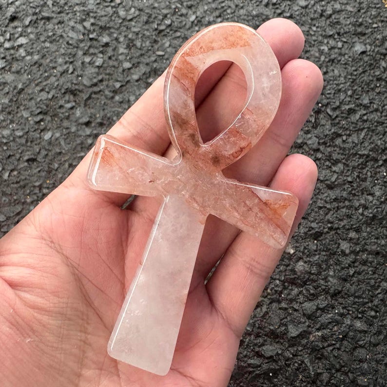 May include: An ankh symbol carved from translucent quartz with visible reddish-brown mineral inclusions throughout the stone. The object features a smooth, polished surface with a rounded top loop and a vertical base, showcasing natural crystalline patterns.