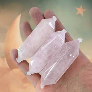 May include: Four rose quartz crystal wands, each with a hexagonal shape and a small white cap on one end. The crystals are a pale pink color and are held in a hand against a soft, dreamy background.
